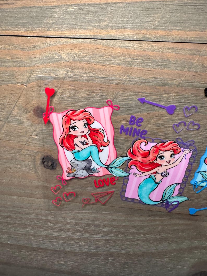 May include: Two die-cut stickers featuring a cartoon mermaid with red hair and a teal tail. One mermaid sits on a rock, the other waves. The stickers include pink striped backgrounds, red and purple hearts, arrows, a paper airplane, and the text Be MINE.