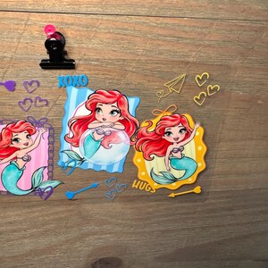 May include: A clear sheet featuring three cartoon mermaid illustrations with red hair and teal tails. The designs include blue and yellow frames, the text XOXO and HUGS, various heart shapes, a paper airplane, and arrows in blue and yellow colors.
