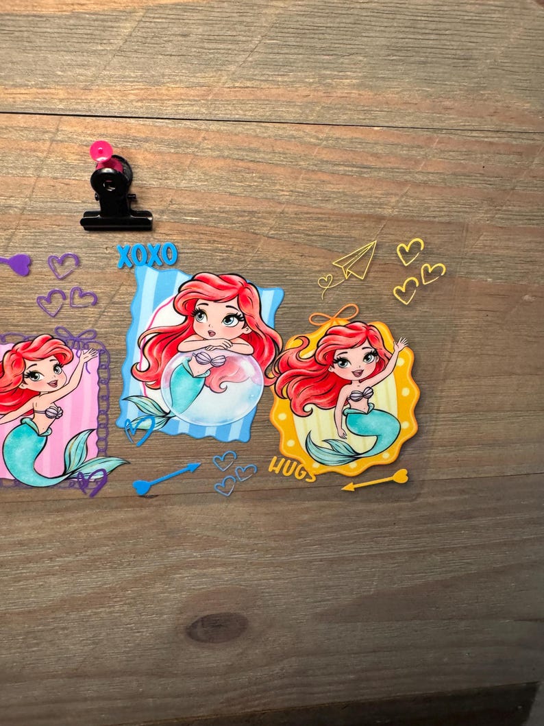 May include: A clear sheet featuring three cartoon mermaid illustrations with red hair and teal tails. The designs include blue and yellow frames, the text XOXO and HUGS, various heart shapes, a paper airplane, and arrows in blue and yellow colors.
