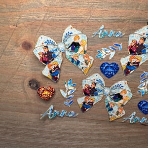 May include: Two hair bows featuring a light blue pattern with various cartoon character illustrations, two blue and orange heart-shaped gems, two blue leaf-shaped accents, and three script-style cutouts of the name Anna in light blue.