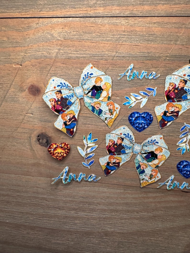 May include: Two hair bows featuring a light blue pattern with various cartoon character illustrations, two blue and orange heart-shaped gems, two blue leaf-shaped accents, and three script-style cutouts of the name Anna in light blue.