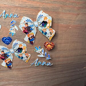May include: Two fabric bows featuring a light blue background with a repeating pattern of cartoon characters, two script cutouts of the name Anna, two leaf-shaped accents, one blue heart-shaped gem, and one orange heart-shaped gem.