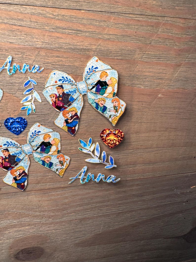May include: Two fabric bows featuring a light blue background with a repeating pattern of cartoon characters, two script cutouts of the name Anna, two leaf-shaped accents, one blue heart-shaped gem, and one orange heart-shaped gem.