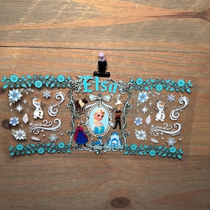 Puede incluir: Una pegatina rectangular transparente con el nombre "Elsa" en turquesa. El dise&ntilde;o incluye personajes y elementos de la pel&iacute;cula, como copos de nieve, flores y un castillo, enmarcados por un borde floral turquesa.