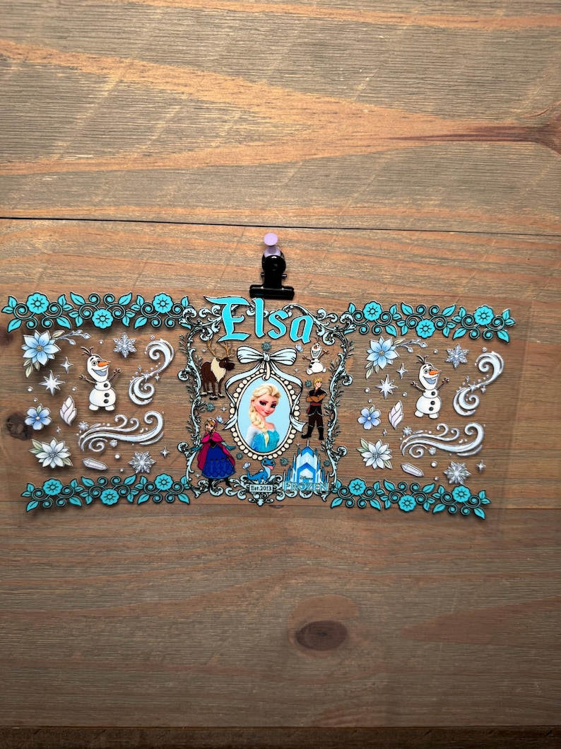 Puede incluir: Una pegatina rectangular transparente con el nombre "Elsa" en turquesa. El dise&ntilde;o incluye personajes y elementos de la pel&iacute;cula, como copos de nieve, flores y un castillo, enmarcados por un borde floral turquesa.