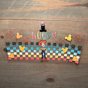 May include: A decorative acrylic wrap featuring a checkered pattern in shades of blue, white, and pink, with the name WOODY in colorful letters, a central Woody character illustration, orange firework accents, and yellow Mickey-shaped balloons.