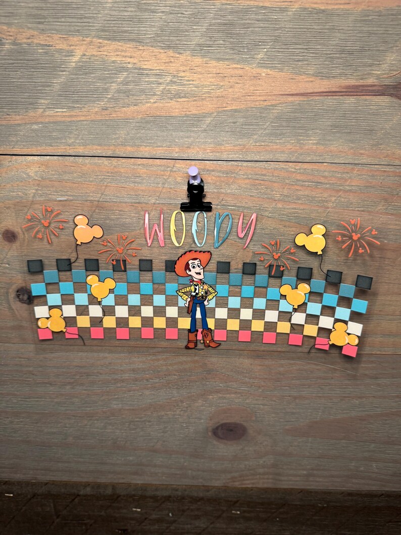 May include: A decorative acrylic wrap featuring a checkered pattern in shades of blue, white, and pink, with the name WOODY in colorful letters, a central Woody character illustration, orange firework accents, and yellow Mickey-shaped balloons.