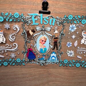 Puede incluir: Una hoja de pegatinas decorativas con personajes de la pel&iacute;cula Frozen. El dise&ntilde;o incluye el nombre "Elsa" en escritura azul, im&aacute;genes de Elsa, Olaf, Kristoff y Anna, con copos de nieve y detalles florales. La hoja de pegatinas est&aacute; sujeta por una pinza negra.
