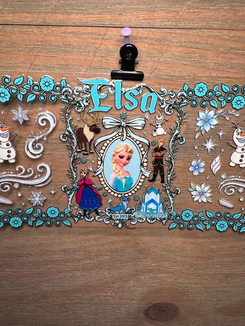 Puede incluir: Una hoja de pegatinas decorativas con personajes de la pel&iacute;cula Frozen. El dise&ntilde;o incluye el nombre "Elsa" en escritura azul, im&aacute;genes de Elsa, Olaf, Kristoff y Anna, con copos de nieve y detalles florales. La hoja de pegatinas est&aacute; sujeta por una pinza negra.