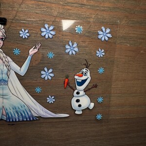K&ouml;nnte beinhalten: Eine transparente Acrylplatte mit einer gedruckten Illustration einer blonden Figur in einem langen blauen Kleid mit Zopf, neben einem kleinen wei&szlig;en Schneemann, der eine orangefarbene Karotte h&auml;lt, umgeben von verschiedenen hellblauen Schneeflocken.