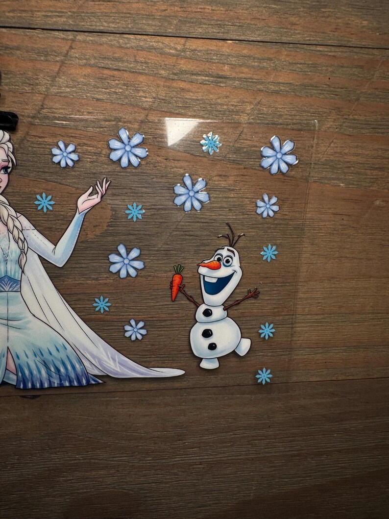 K&ouml;nnte beinhalten: Eine transparente Acrylplatte mit einer gedruckten Illustration einer blonden Figur in einem langen blauen Kleid mit Zopf, neben einem kleinen wei&szlig;en Schneemann, der eine orangefarbene Karotte h&auml;lt, umgeben von verschiedenen hellblauen Schneeflocken.
