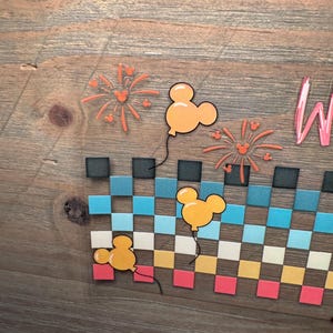 May include: A wooden sign featuring a checkered pattern in rows of black, teal, light blue, white, yellow, and pink squares. Three yellow mouse-shaped balloons and two orange firework designs with small mouse silhouettes are displayed above.