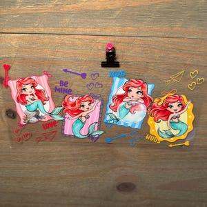 May include: A clear sheet featuring four cartoon mermaid illustrations with red hair and teal tails, framed in pink, purple, blue, and yellow. The design includes text reading Be MINE, XOXO, and HUGS, alongside various heart and arrow graphics.