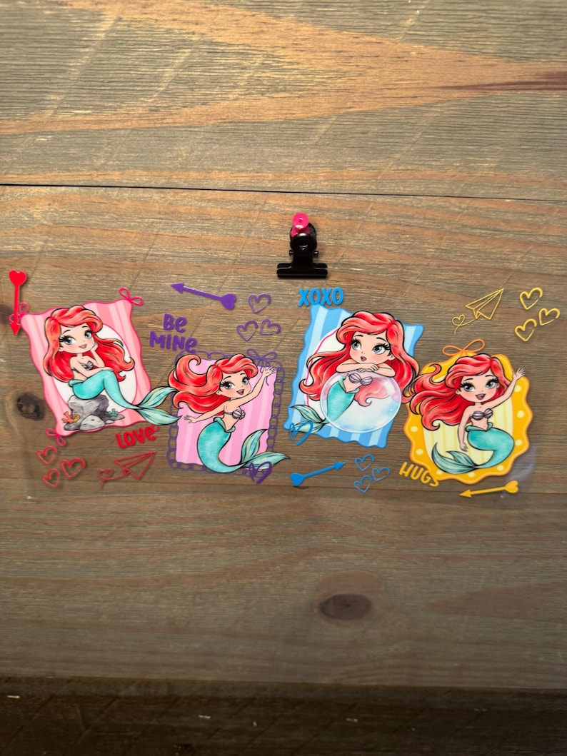 May include: A clear sheet featuring four cartoon mermaid illustrations with red hair and teal tails, framed in pink, purple, blue, and yellow. The design includes text reading Be MINE, XOXO, and HUGS, alongside various heart and arrow graphics.