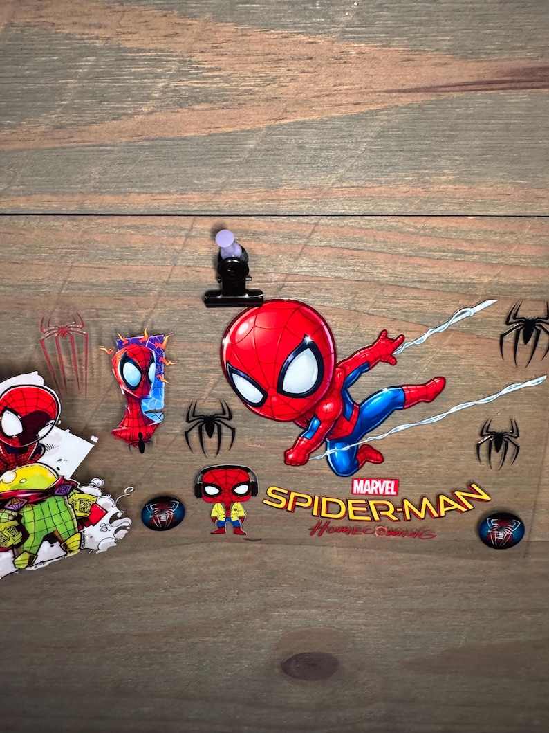 K&ouml;nnte beinhalten: Eine Sammlung von Spider-Man-Aufklebern und Grafiken. Das Hauptbild zeigt einen Cartoon-Spider-Man in einer dynamischen Pose mit den Worten "Marvel Spider-Man Homecoming". Zus&auml;tzliche Aufkleber zeigen Spider-Man-Logos und andere Charakterdesigns.