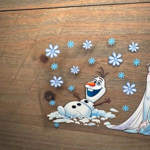 K&ouml;nnte beinhalten: Ein transparenter Aufkleber mit der Figur Olaf aus Die Eisk&ouml;nigin, dargestellt mit einer leuchtend orangefarbenen Karottennase, gro&szlig;en Augen und einem fr&ouml;hlichen Ausdruck, umgeben von zahlreichen hellblauen Schneeflocken-Illustrationen.