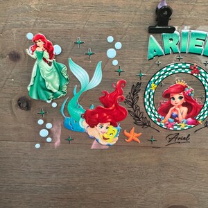 Pu&ograve; includere: Adesivi a tema Ariel, la Sirenetta, in varie pose. Un adesivo mostra Ariel con un abito verde, un altro con la sua coda e un terzo all'interno di una cornice circolare. Gli adesivi includono stelle, bolle e il nome "Ariel".