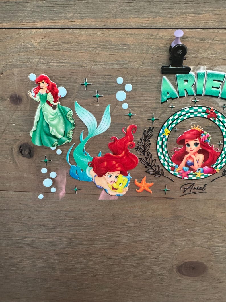 Pu&ograve; includere: Adesivi a tema Ariel, la Sirenetta, in varie pose. Un adesivo mostra Ariel con un abito verde, un altro con la sua coda e un terzo all'interno di una cornice circolare. Gli adesivi includono stelle, bolle e il nome "Ariel".
