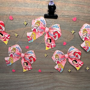 May include: Four fabric hair bows featuring a repeating pattern of cartoon princesses including Cinderella, Aurora, and Ariel on a pink background with blue leaf accents. The bows are arranged with small pink heart and gold star confetti pieces.