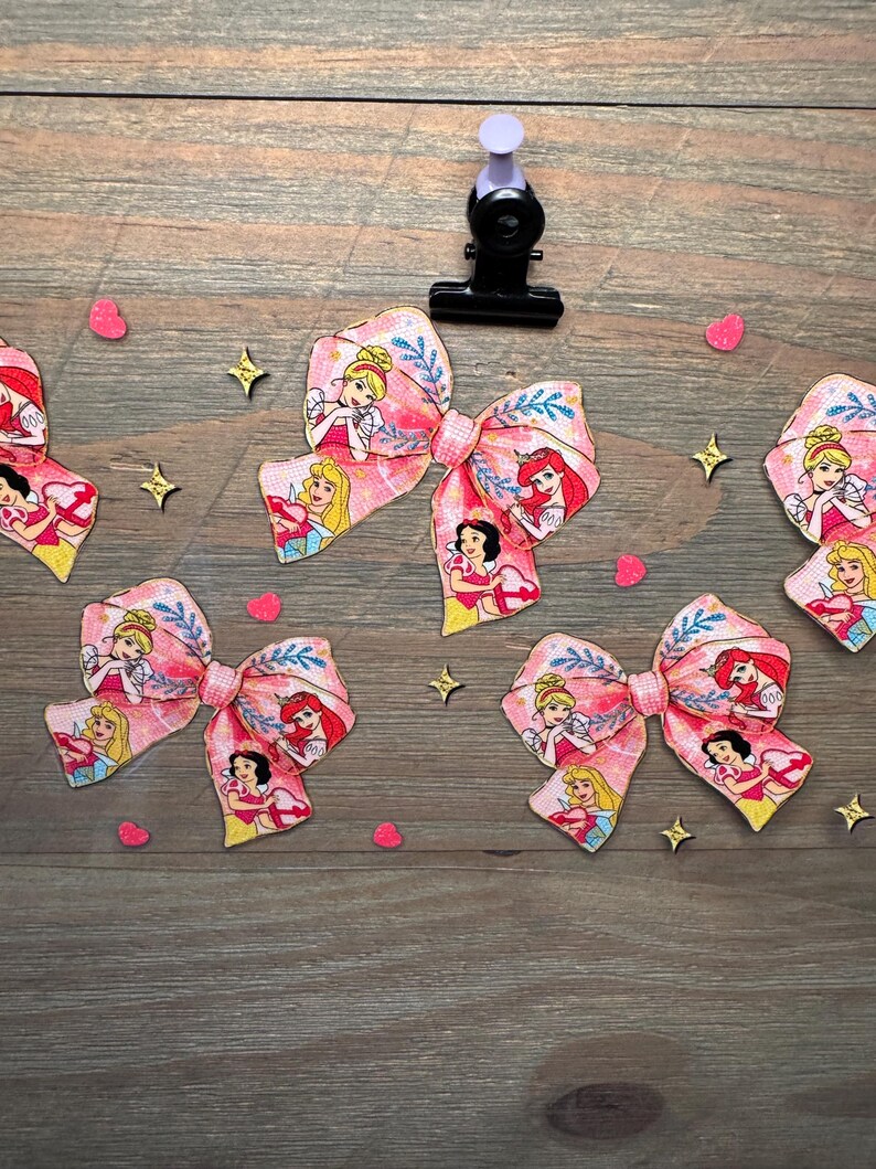 May include: Four fabric hair bows featuring a repeating pattern of cartoon princesses including Cinderella, Aurora, and Ariel on a pink background with blue leaf accents. The bows are arranged with small pink heart and gold star confetti pieces.