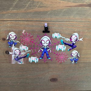 May include: A collection of Spider-Gwen character stickers in various poses. The stickers feature a white and pink color scheme with spiderwebs and spider graphics. The background is a wooden surface.