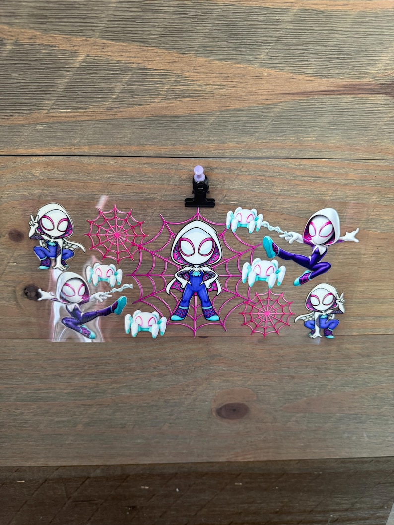 May include: A collection of Spider-Gwen character stickers in various poses. The stickers feature a white and pink color scheme with spiderwebs and spider graphics. The background is a wooden surface.
