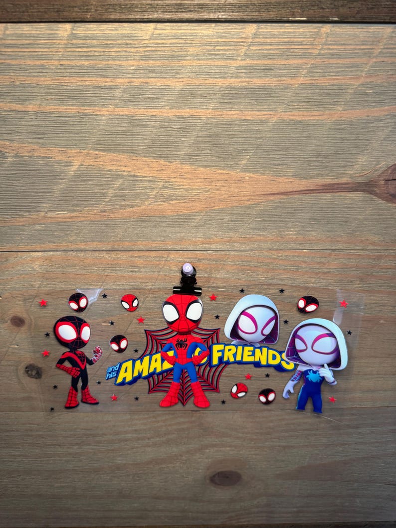 May include: A clear transfer sheet featuring cartoon characters from Spidey and His Amazing Friends, including Miles Morales, Peter Parker, and Ghost-Spider, with red spider web accents, small black stars, and the yellow text "and his AMAZING FRIENDS."