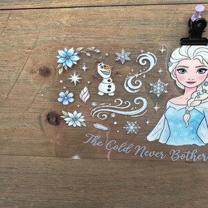 May include: A clear sticker sheet featuring Elsa from Frozen, snowflakes, flowers, and Olaf. The text "The Cold Never Bothered Me Anyway" is written in a cursive font. The sticker sheet is held in place by a black clip with a purple top.