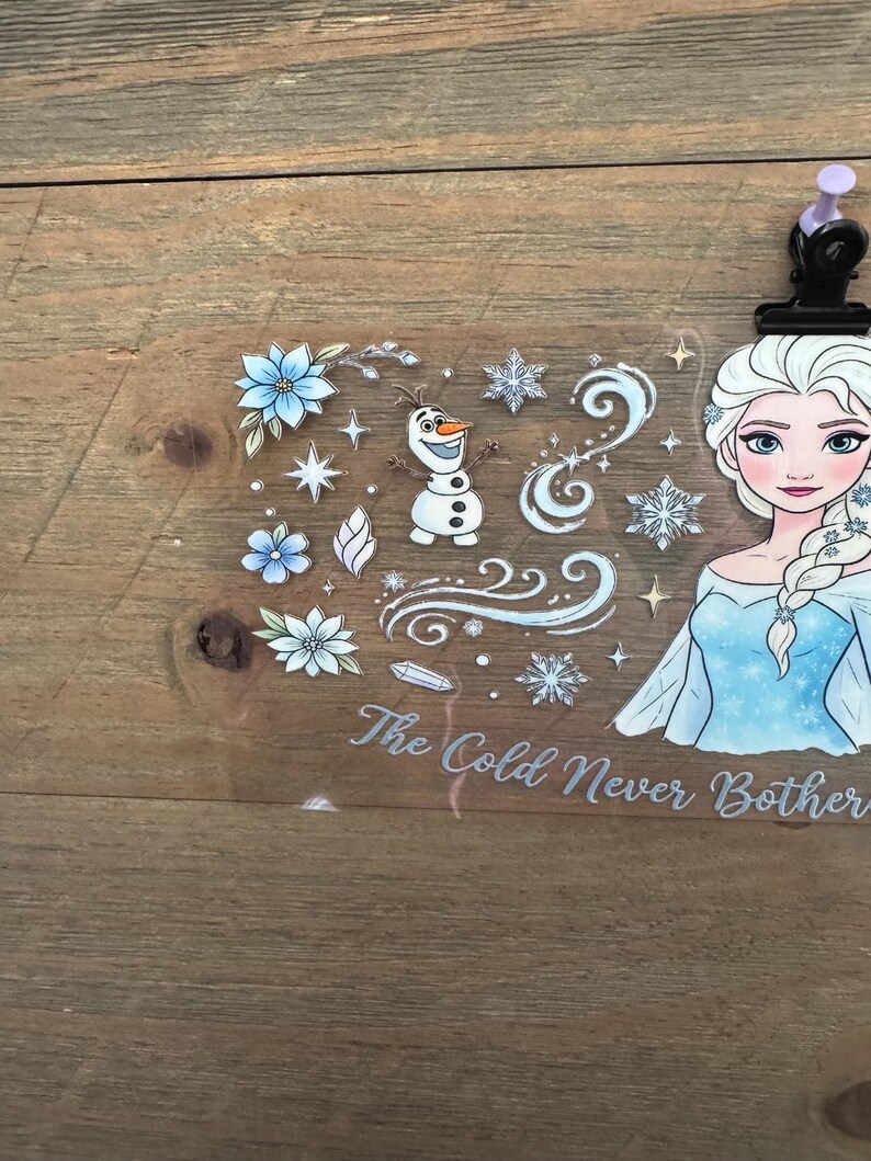 May include: A clear sticker sheet featuring Elsa from Frozen, snowflakes, flowers, and Olaf. The text "The Cold Never Bothered Me Anyway" is written in a cursive font. The sticker sheet is held in place by a black clip with a purple top.