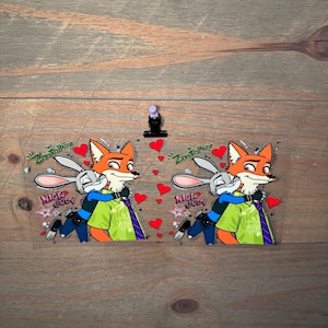 May include: Two clear acrylic charms featuring Nick Wilde and Judy Hopps from Zootopia. The fox and bunny characters are hugging, surrounded by red hearts. Text includes "Zootopia" and "Nick & Judy".