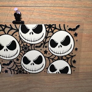 May include: A clear, rectangular Halloween-themed decoration featuring multiple white Jack Skellington faces with black eyes and mouths. The design includes black spiderwebs, bats, and stars. A black clip with a purple top holds the decoration.