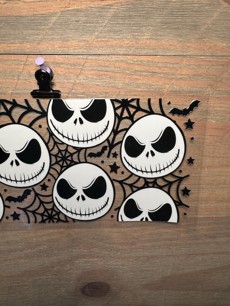 May include: A clear, rectangular Halloween-themed decoration featuring multiple white Jack Skellington faces with black eyes and mouths. The design includes black spiderwebs, bats, and stars. A black clip with a purple top holds the decoration.