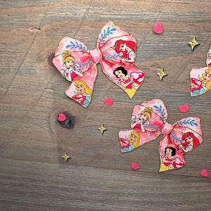 May include: Two fabric hair bows featuring a pink background with a repeating pattern of cartoon princesses, blue floral accents, and a small checkered center knot. The bows are decorated with small red heart and gold star confetti pieces.