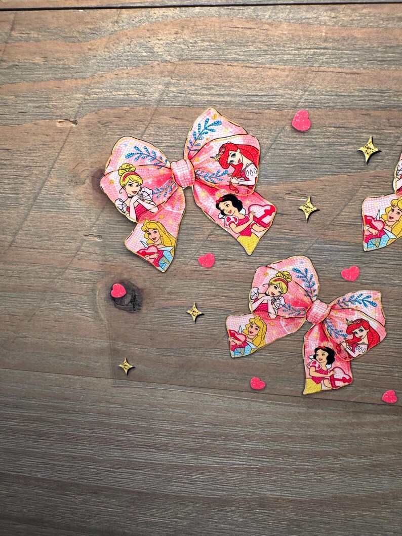 May include: Two fabric hair bows featuring a pink background with a repeating pattern of cartoon princesses, blue floral accents, and a small checkered center knot. The bows are decorated with small red heart and gold star confetti pieces.