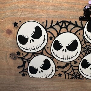 May include: Black and white Halloween-themed decoration featuring multiple Jack Skellington faces. The design includes spiderwebs, bats, and stars. The decoration is placed on a wooden surface.