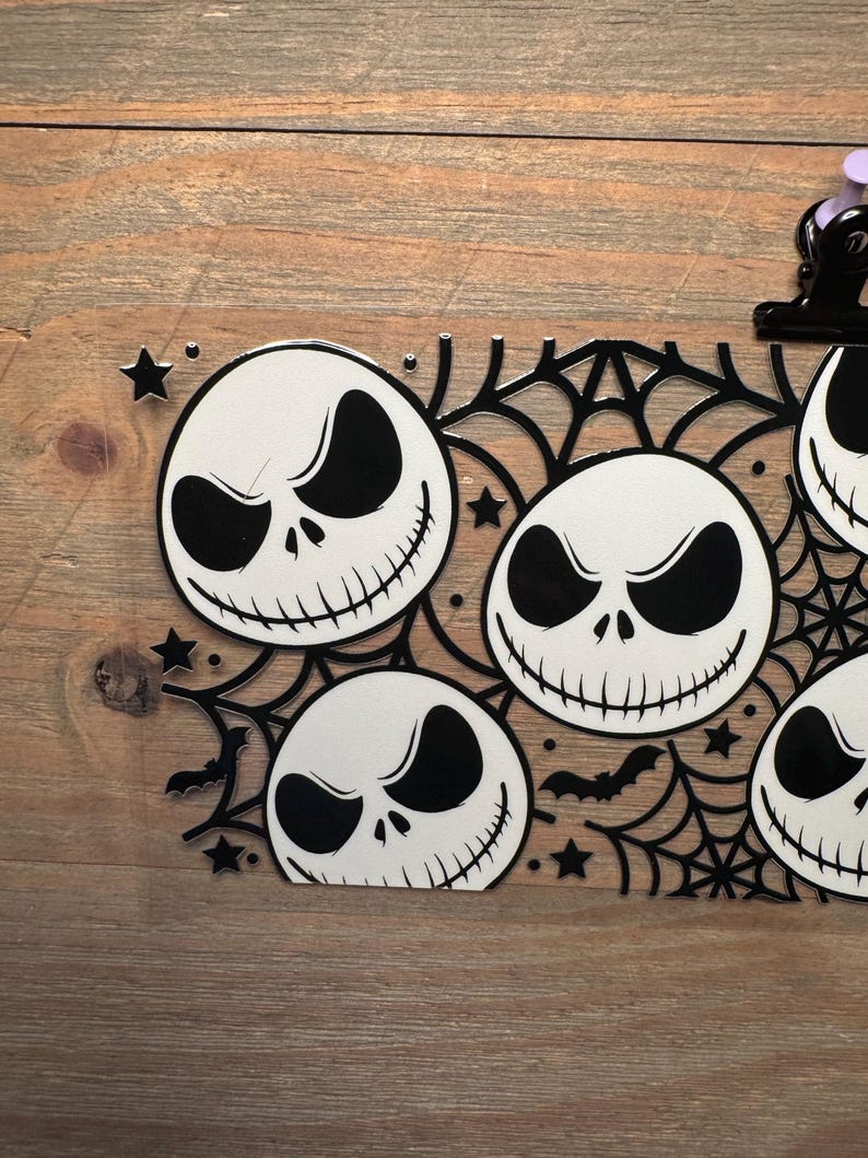 May include: Black and white Halloween-themed decoration featuring multiple Jack Skellington faces. The design includes spiderwebs, bats, and stars. The decoration is placed on a wooden surface.