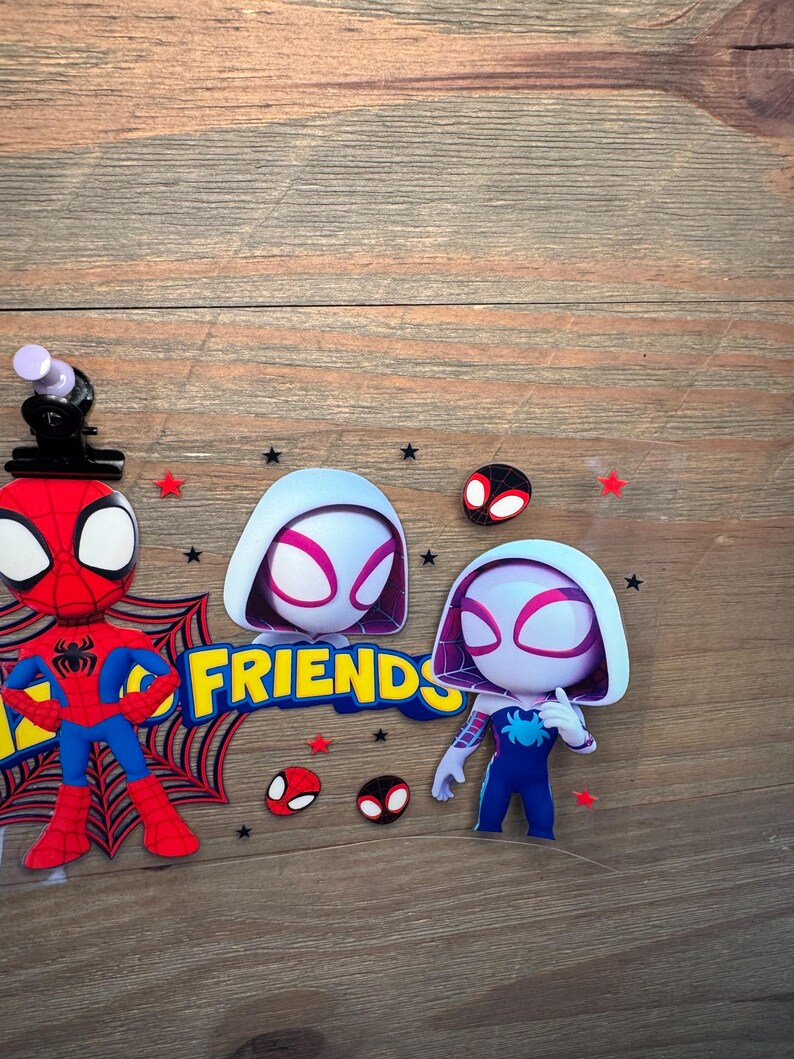 May include: A clear plastic sheet featuring cartoon character stickers including a red and blue spider-themed hero, a white and purple hooded character, and the yellow text FRIENDS. The design includes small red stars, black stars, and spider mask icons.