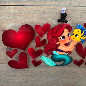May include: Ariel and Flounder illustration with red hearts. Ariel has red hair, a blue top, and a teal tail. Flounder is yellow and blue. The hearts are various sizes and shades of red. The image is on a wooden surface.