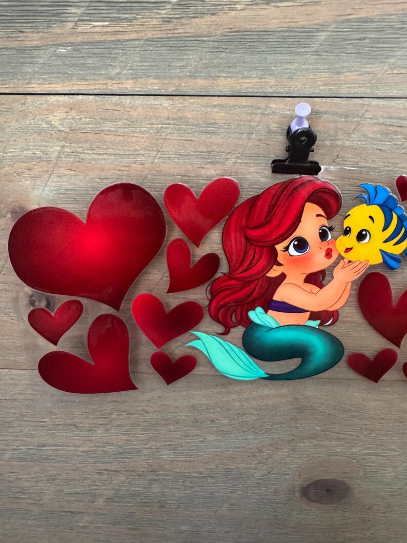 May include: Ariel and Flounder illustration with red hearts. Ariel has red hair, a blue top, and a teal tail. Flounder is yellow and blue. The hearts are various sizes and shades of red. The image is on a wooden surface.