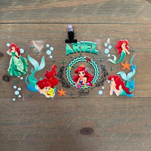 Pu&ograve; includere: Un foglio di plastica trasparente con Ariel de La Sirenetta. Il design include pi&ugrave; immagini di Ariel con capelli rossi e coda verde, circondata da bolle e stelle marine. La parola "ARIEL" &egrave; ben visibile.