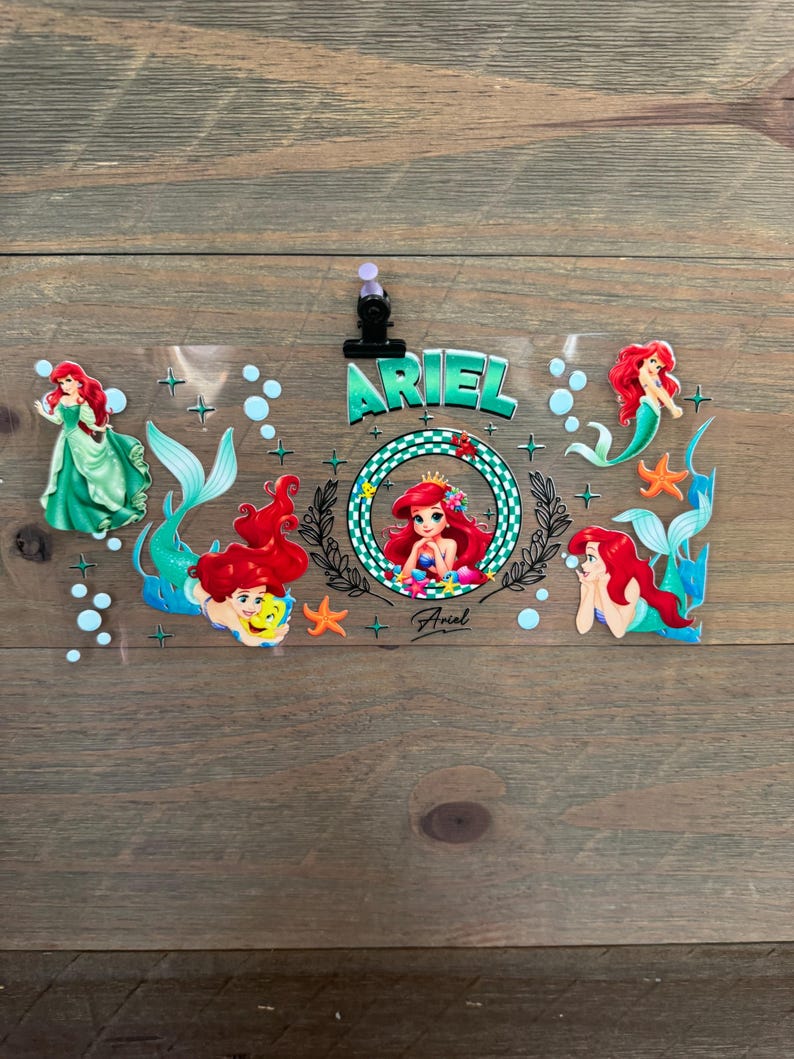 Pu&ograve; includere: Un foglio di plastica trasparente con Ariel de La Sirenetta. Il design include pi&ugrave; immagini di Ariel con capelli rossi e coda verde, circondata da bolle e stelle marine. La parola "ARIEL" &egrave; ben visibile.