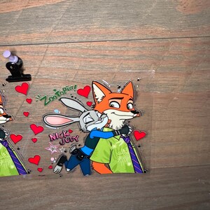 May include: A clear, rectangular sticker featuring the characters Nick Wilde and Judy Hopps from Zootopia. Nick, an orange fox, is embracing Judy, a gray rabbit. The sticker includes the text "Zootopia!" and "Nick Judy" with red heart accents.