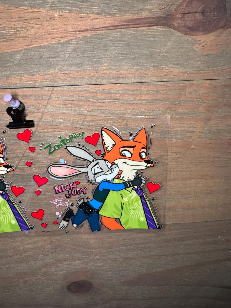 May include: A clear, rectangular sticker featuring the characters Nick Wilde and Judy Hopps from Zootopia. Nick, an orange fox, is embracing Judy, a gray rabbit. The sticker includes the text "Zootopia!" and "Nick Judy" with red heart accents.