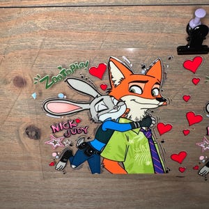 May include: A clear sticker featuring the characters Nick Wilde and Judy Hopps from Zootopia. Nick, an orange fox, hugs Judy, a gray bunny. The sticker includes the text "Zootopia!" and "Nick Judy", along with red heart and star accents.
