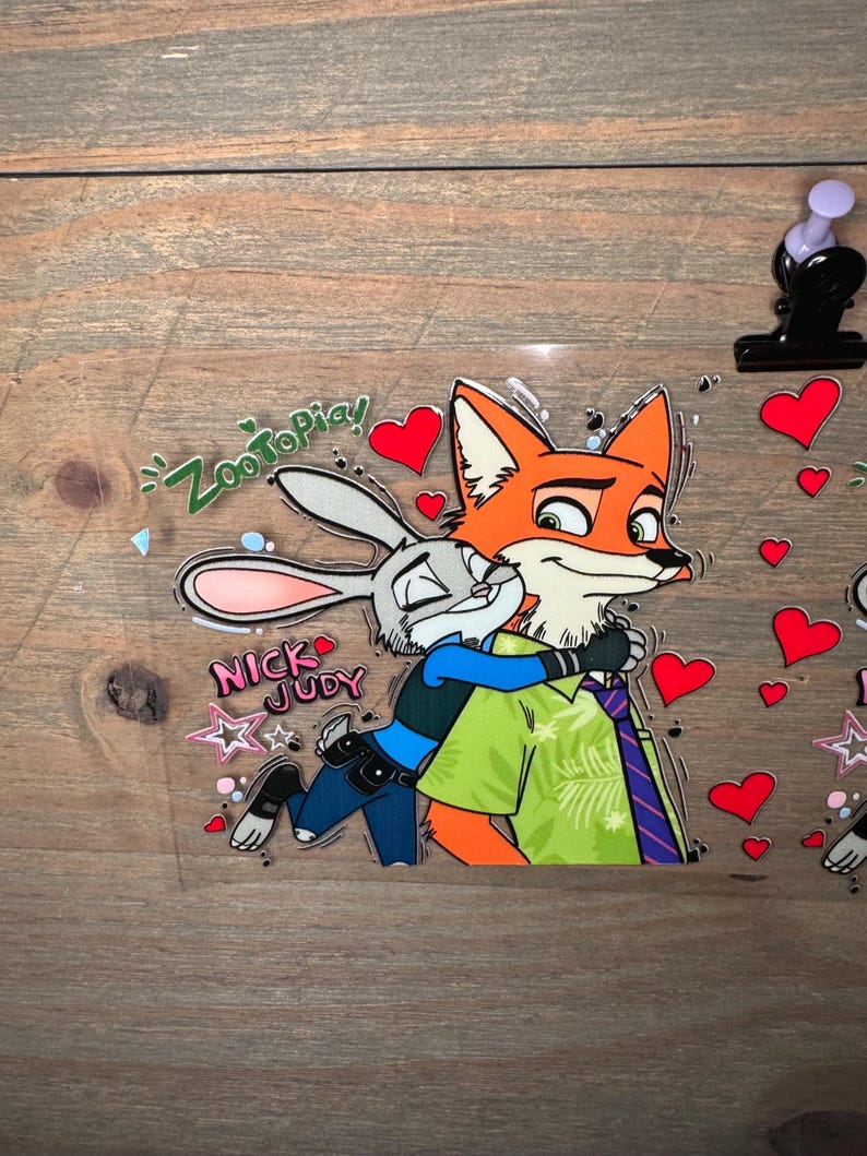 May include: A clear sticker featuring the characters Nick Wilde and Judy Hopps from Zootopia. Nick, an orange fox, hugs Judy, a gray bunny. The sticker includes the text "Zootopia!" and "Nick Judy", along with red heart and star accents.