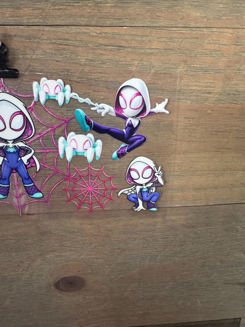 May include: A collection of Spider-Gwen character cutouts in various poses. The figures are primarily purple, white, and blue, with spider web and spider accents. One figure is mid-air, attached to a web.