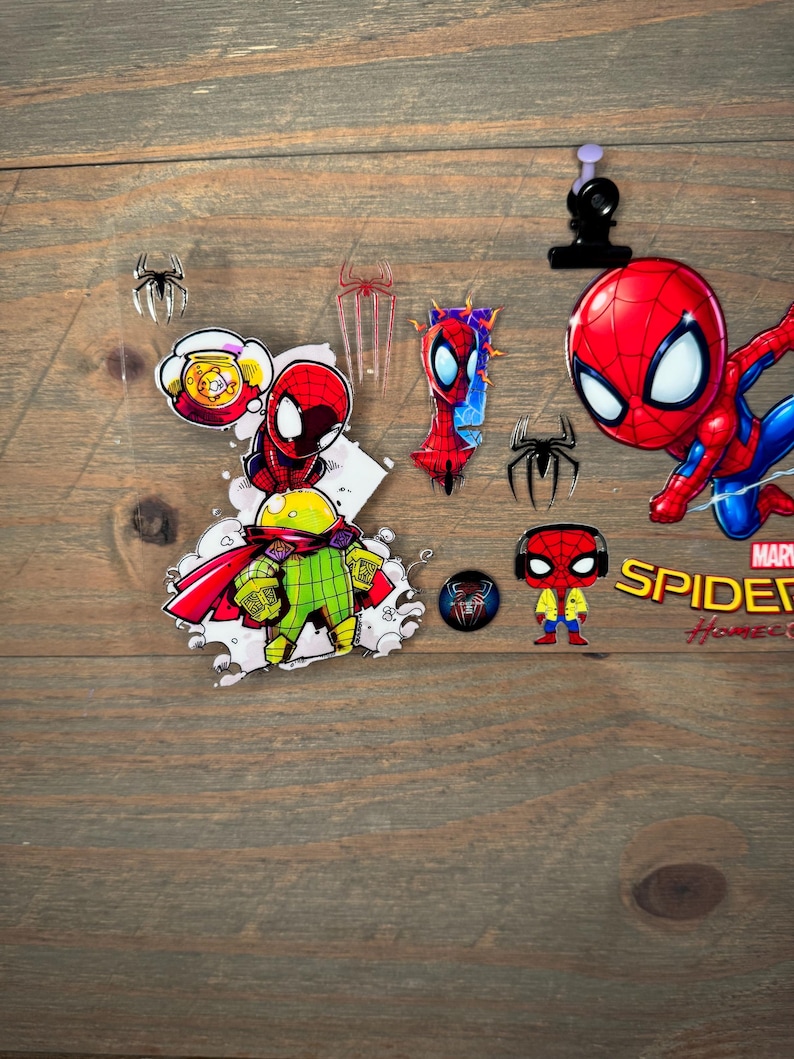 K&ouml;nnte beinhalten: Eine Sammlung von Spider-Man-Artikeln, darunter Aufkleber, Anstecknadeln und eine kleine Figur. Die Artikel zeigen den Superhelden in seinem charakteristischen rot-blauen Anzug. Der Hintergrund ist eine Holzoberfl&auml;che.