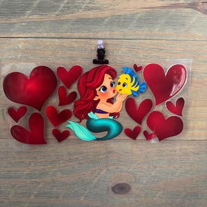 May include: A clear plastic clip featuring a cartoon mermaid with red hair and a teal tail, kissing a yellow and blue fish. Red hearts surround the characters, creating a romantic theme. The clip is attached to a wooden surface.
