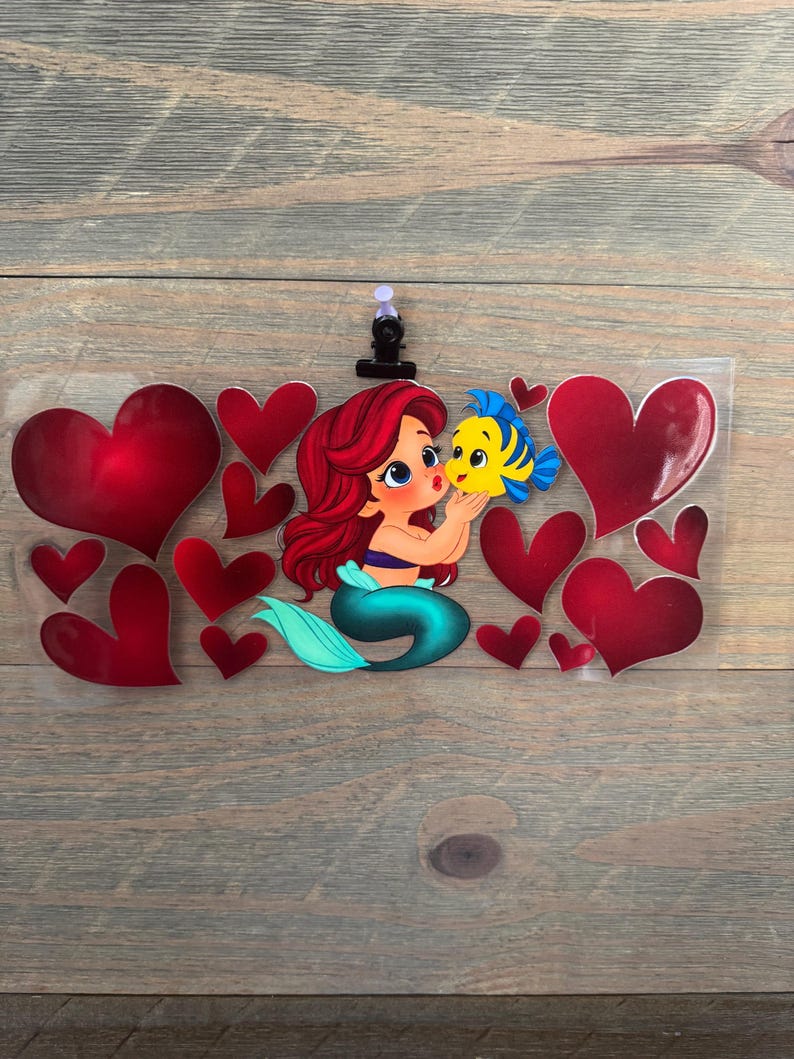 May include: A clear plastic clip featuring a cartoon mermaid with red hair and a teal tail, kissing a yellow and blue fish. Red hearts surround the characters, creating a romantic theme. The clip is attached to a wooden surface.
