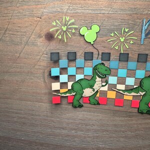 Puede incluir: Una manualidad de papel en capas que presenta un dinosaurio de dibujos animados verde frente a un patr&oacute;n de cuadros que cambia de negro a azul, beige, naranja y rojo. Sobre el dinosaurio hay fuegos artificiales verde claro y un globo con forma de Mickey.